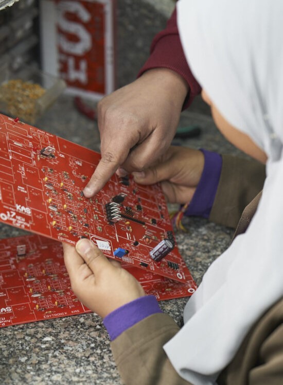a girl holds a circuit board while a hand shows something on it