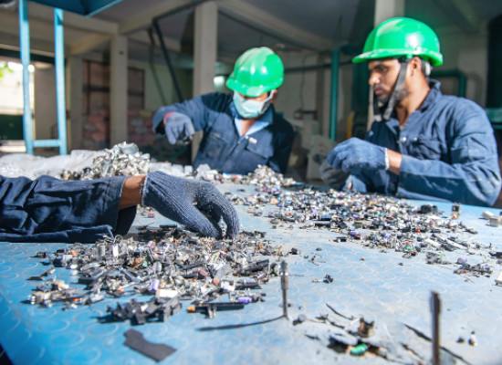 Electronic waste on a shop board with two workers in the background selecting parts for recycling and a hand of a third worker coming from the left side, also selecting parts.