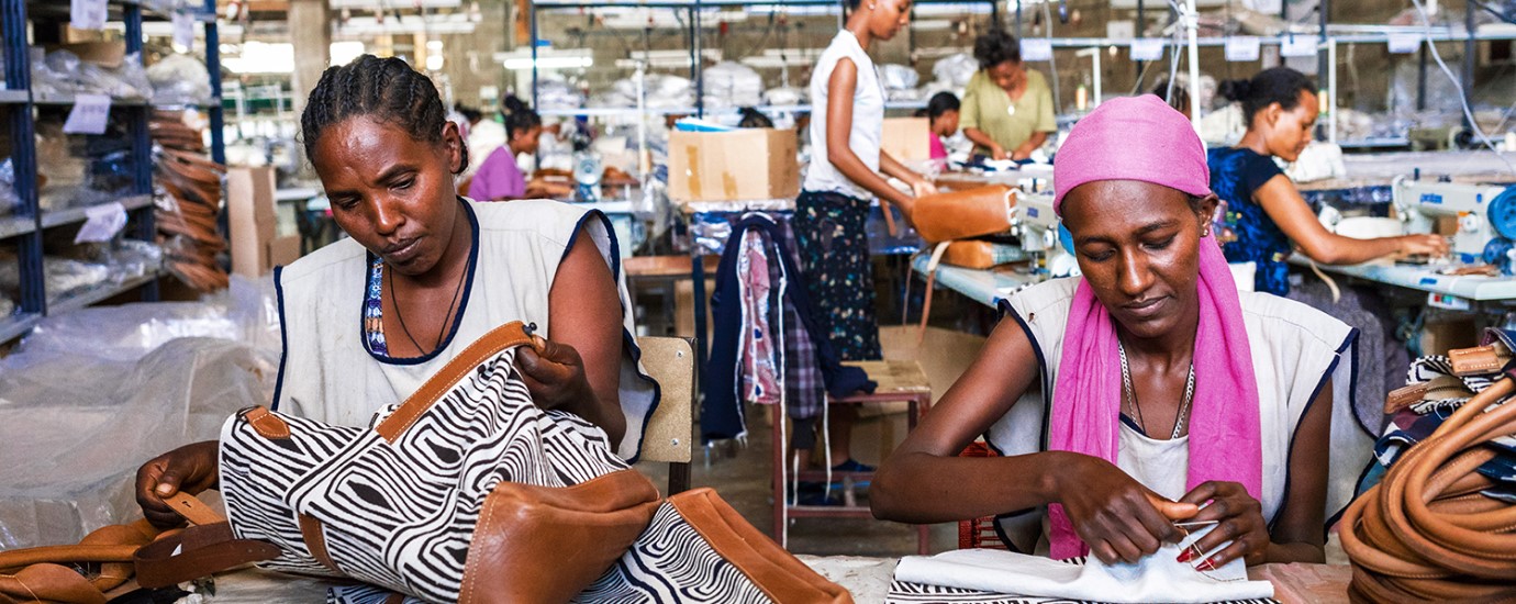 Two women sewing leather bags in a factory. Women in the background, sewing.