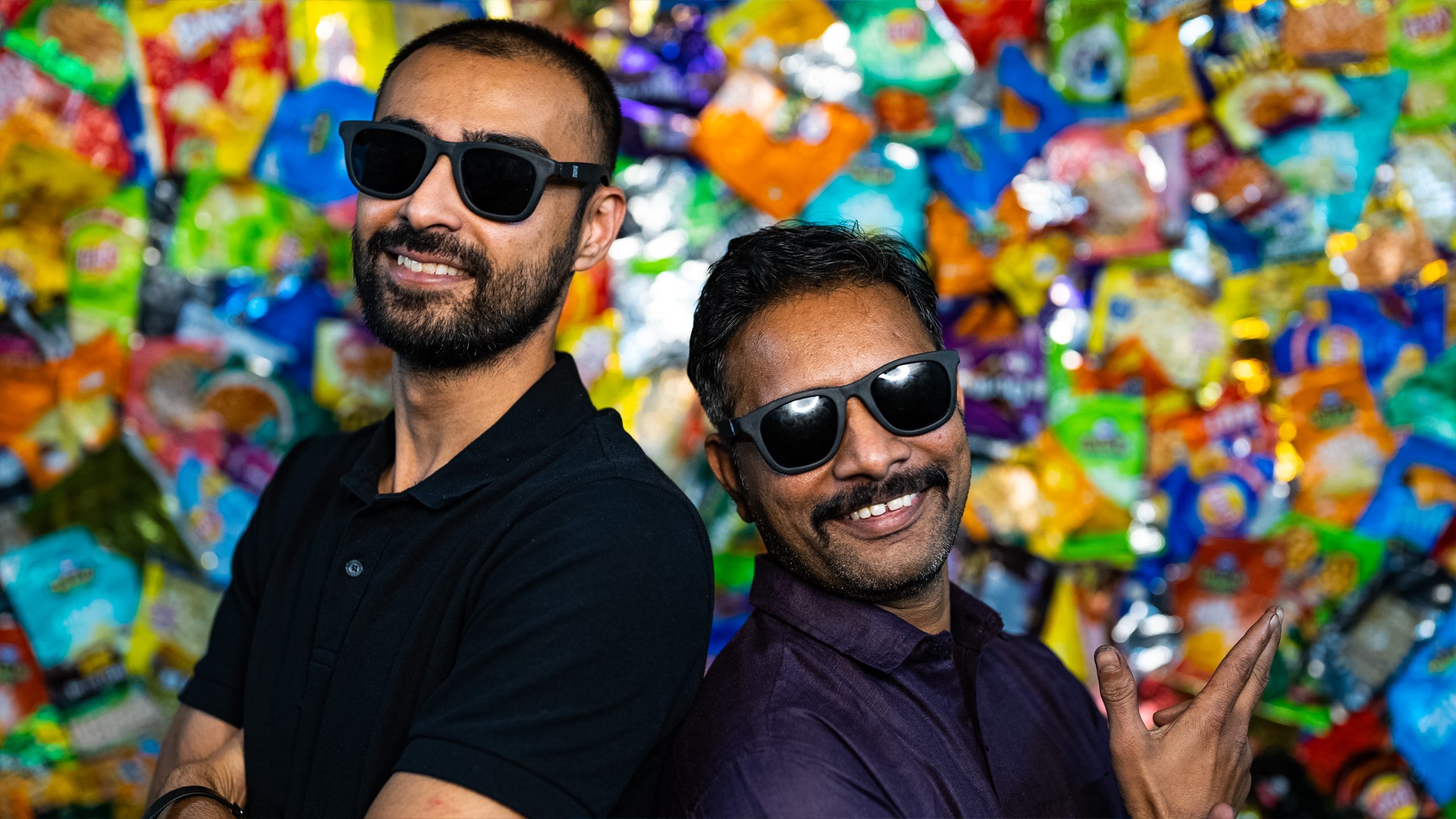 Two Indian men with sunglasses in front of a wall decoratded with colourfull chips packets