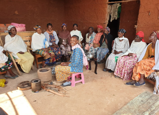 10+ african women sitting in a circle on chairs in a clay house, a woman in the middle of the room, smiling at camera. Stove and parts of it are laying before her
