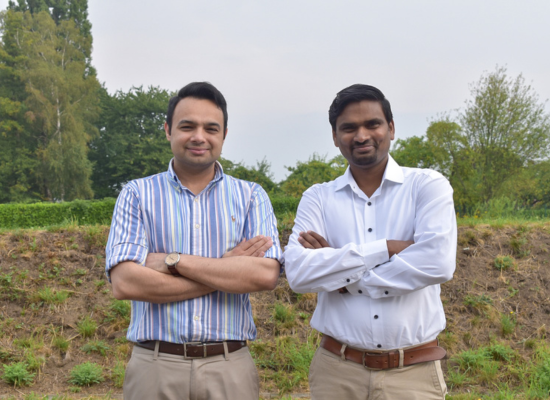 two indian men smiling at camera, arms corossed, field/ hill in background