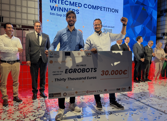 Two men with short beard are holding an oversized award check of 30000 €; one man raises his fist in the air in a triumphal manner