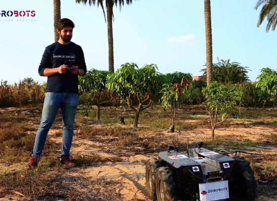 Man with beard is holding controller, standing next to a robot with four thick wheels on a field. A couple of palms and bushes in the background