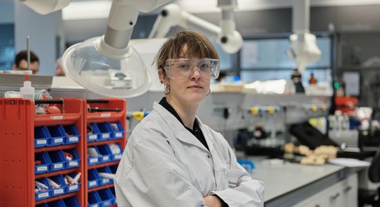 A woman wearing a lab coat and protective glasses poses in a laboratory.