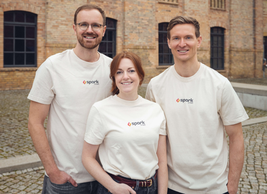 Three people (two man & one woman) in white T-shirts with company logo on it smiling into the camera. Red brick building in the background.