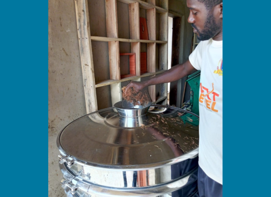 African man putting a handfull of coconut husks into metall reaction container.